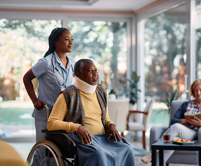 Injured African American senior man in wheelchair being pushed by young nurse in nursing home.